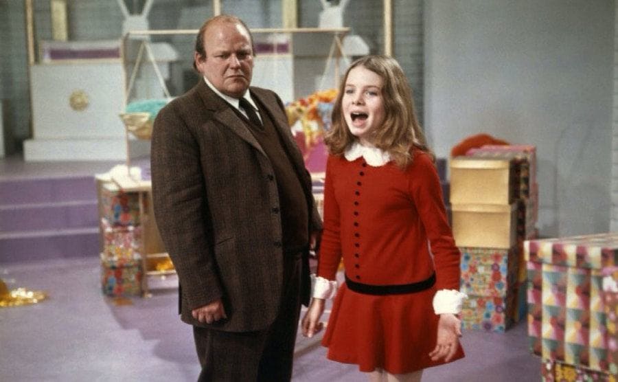 Veruca singing next to her father in a room full of boxes and ribbon 
