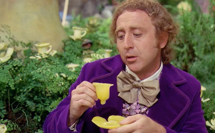Willy Wonka sitting in front of bushes with a yellow tea cup 