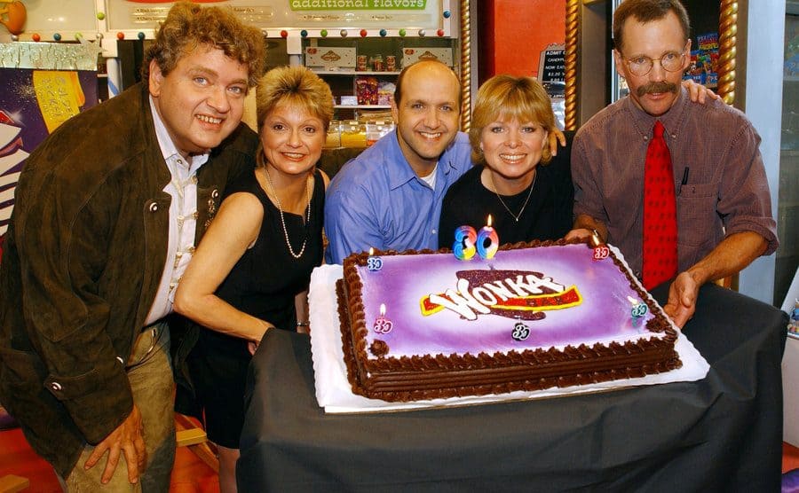 The five kids from the cast of Willy Wonka and the Chocolate Factory posing around a cake with a Wonka bar logo and candles that spell out 30 lit up 