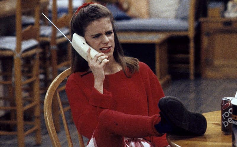 Andrea Barber as Kimmy Gibbler sitting at the kitchen table talking on the phone with her feet up 
