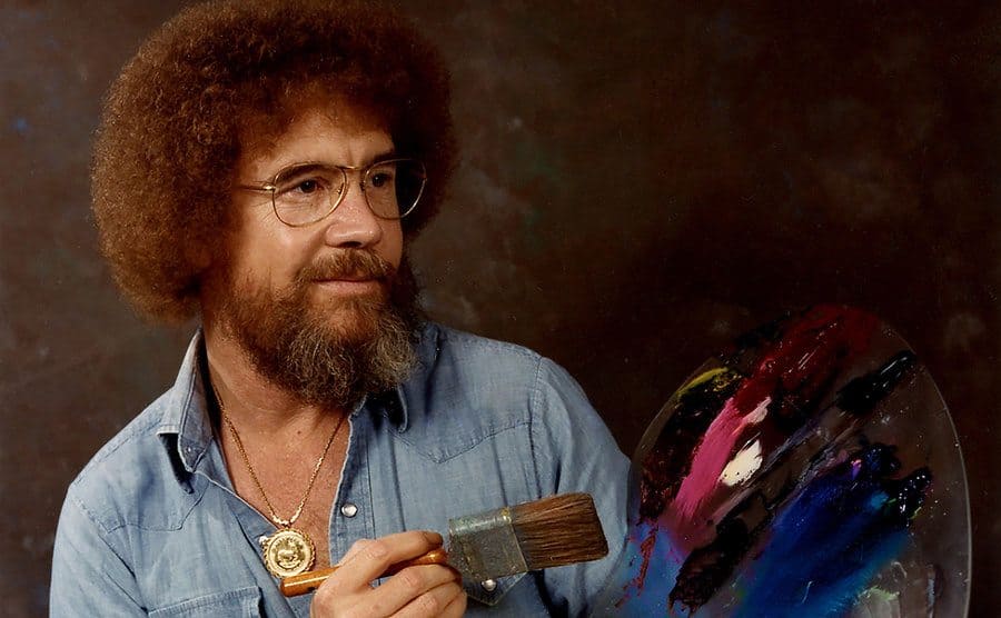 Bob Ross is posing with his paintbrush and palette. 