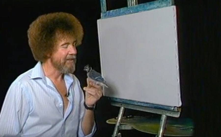 Bob Ross on set talking to a bird as he holds it in his hand. 