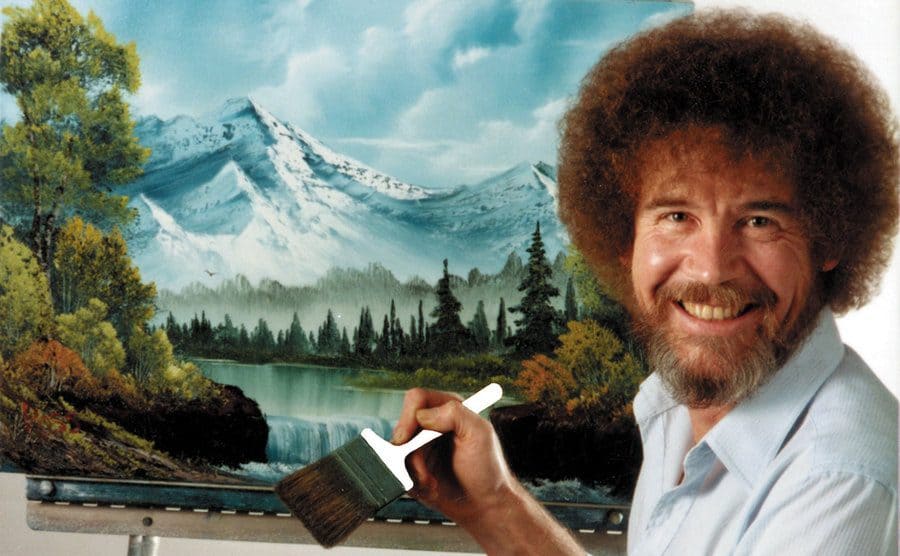 Bob Ross is smiling in a promotional photo in front of one of his paintings. 