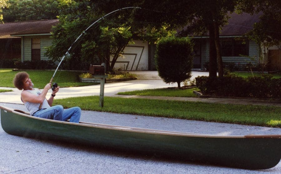 Bob Ross is fishing from his boat, which is placed in the middle of the street. 