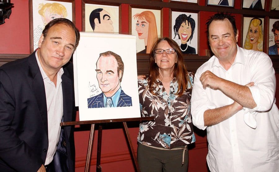 Jim Belushi, Judy Belushi, and Dan Aykroyd posing around a caricature 