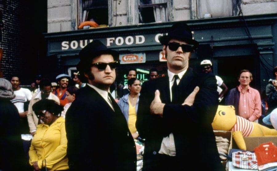 John Belushi and Dan Aykroyd on the set of the Blues Brothers wearing black suits and hats 