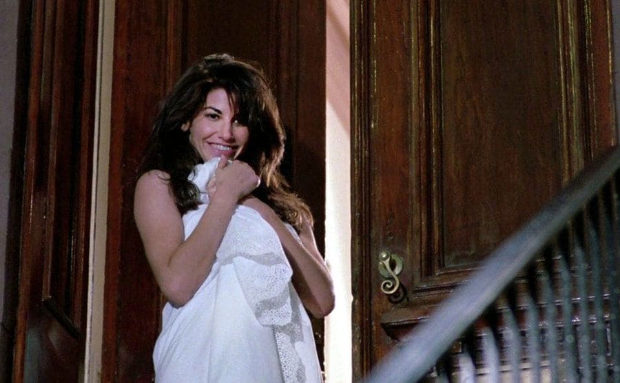 Gina Gershon is standing next to the door with a smile on her face wrapped in a white blanket. 