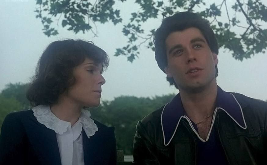 Travolta and Gorney are sitting on a bench by the bridge. 