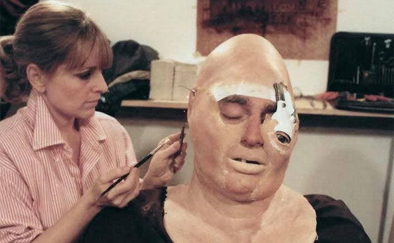 John Matuszak is sitting in the makeup chair, having his prosthetics placed on his face. 