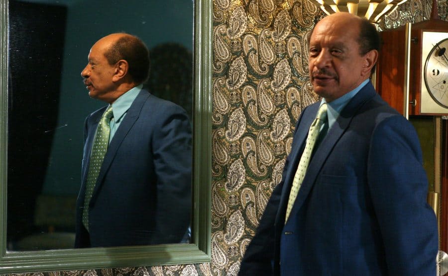 Sherman Hemsley is passing next to an indoors mirror.