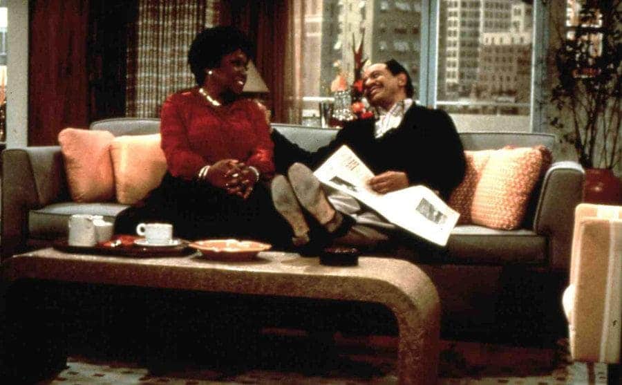 Movie still of Sherman Hemsley and Isabel Sanford sitting and relaxing in the living room behind a small table while talking and reading the newspaper.