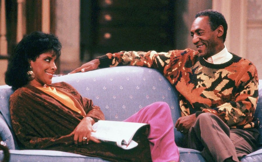 Bill Cosby, as Heathcliff Huxtable, sitting on the couch beside Phylicia Rashad, as Clair Huxtable. 