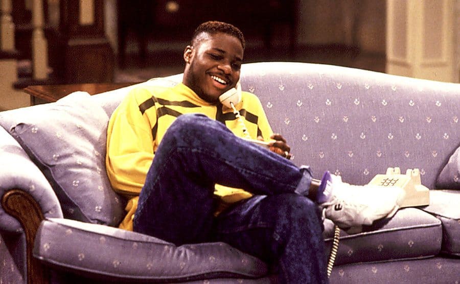 Theo Huxtable, played by Malcolm-Jamal Warner, is sitting on the couch while talking on the phone. 