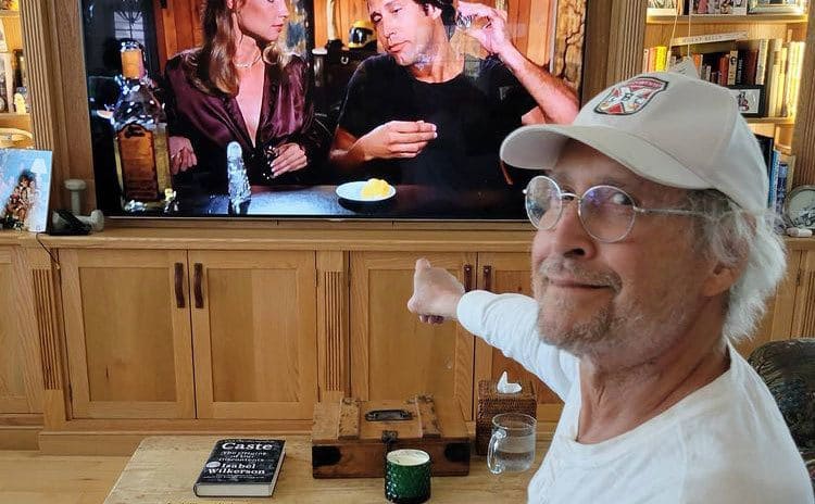 Chevy Chase sits in his house, pointing at himself on the television screen.