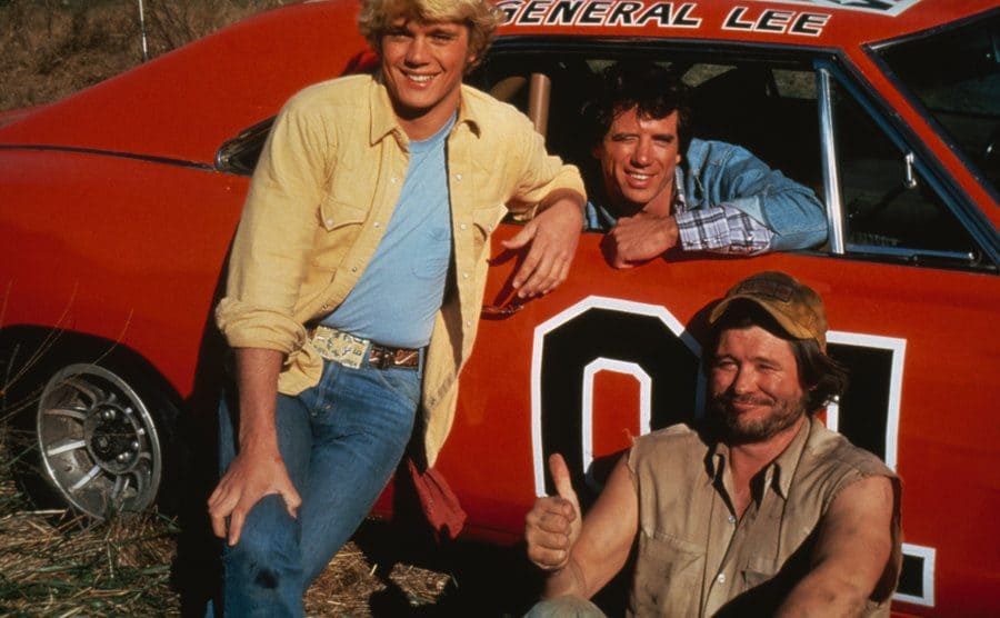 John Schneider, Tom Wopat, and Ben Jones are posing with the General Lee, a 1969 Dodge Charger prominently featured in the series.
