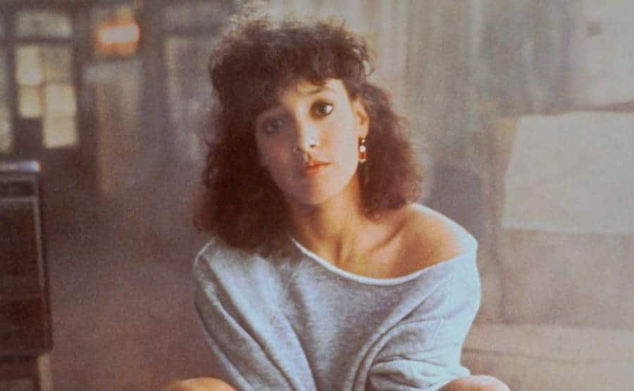Jennifer Beals is posing, dressed in the famous off-the-shoulder sweatshirt from Flashdance.