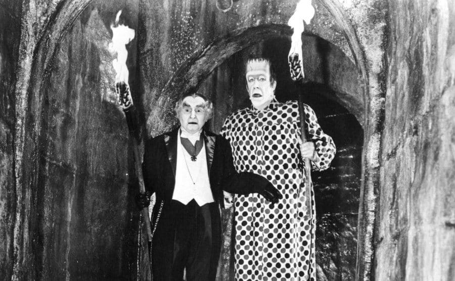 A still of Grandpa and Herman Munster holding torches in a foggy corridor.