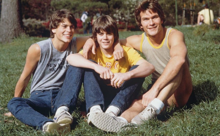 Rob Lowe, C. Thomas Howell, and Patrick Swayze are sitting on the grass.