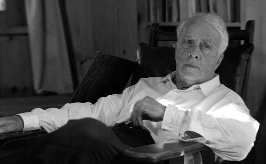Poet Robert Frost is reclining in a wooden chair wearing a reflective expression on his face.