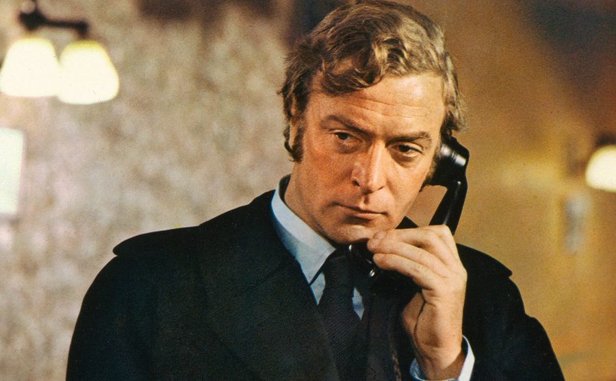 Michael Caine wearing a black raincoat and holding a black telephone receiver.