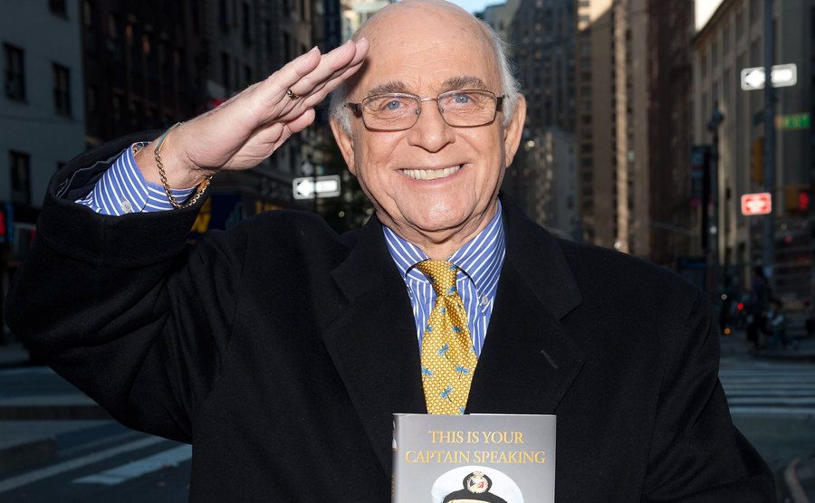 Gavin MacLeod holding up a book and saluting.