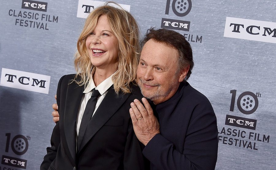 Billy Crystal crouches behind Meg Ryan while posing for the press at an event. 