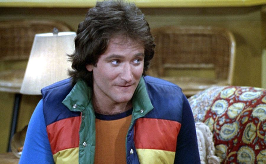 Robin Williams, as Mork, sits in his puffer vest on the couch. 