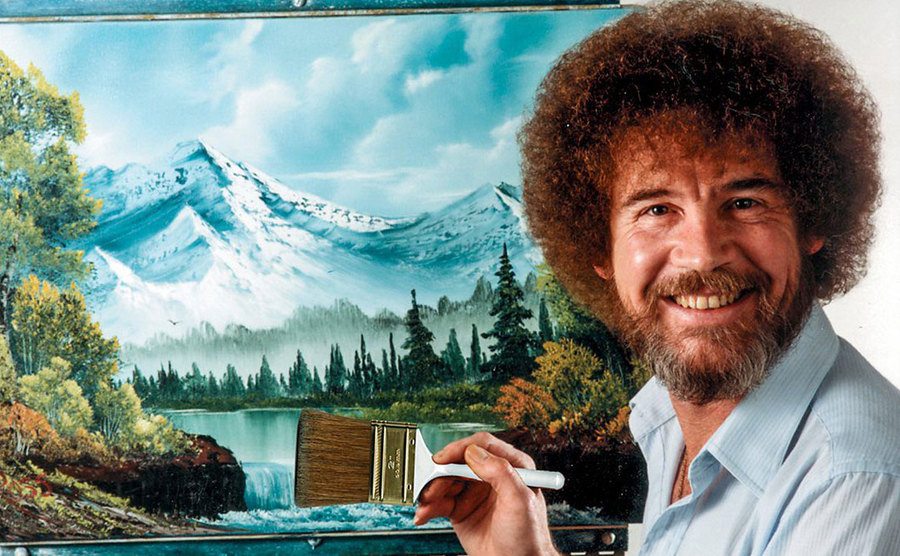 Bob Ross poses for a photo while working on a painting. 